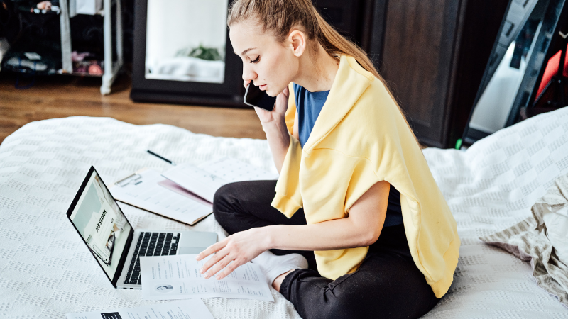 A woman sitting on a bed, using a laptop and phone for work or leisure.