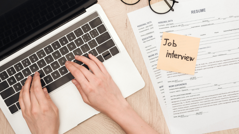 Person working on a laptop with their resume beside them and a note saying job interview.